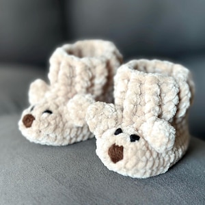 May include: A pair of beige crocheted baby booties with bear faces. The booties have brown felt noses and black felt eyes.