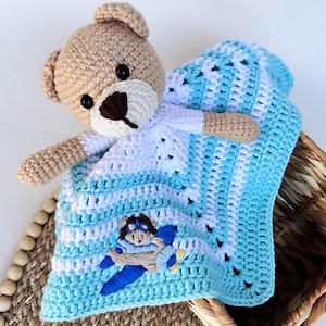 May include: A crocheted baby blanket with a brown teddy bear attached. The blanket is blue and white with a stitched design of a bear in a plane.