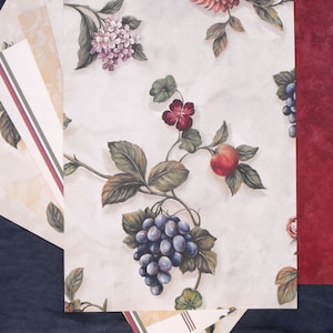 May include: A collection of decorative papers featuring floral and fruit designs. The main paper has a cream background with illustrations of grapes, apples, and flowers in shades of purple, red, and green. A deep red paper and other patterned papers are also included.