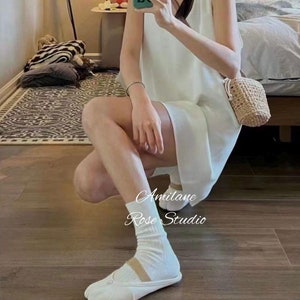 May include: A woman wearing a white sleeveless dress and white socks with white tabi shoes. She is holding a small woven bag. The text "Amilane Rose Studio" is visible in the image.