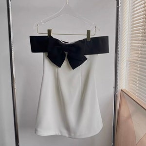 Könnte beinhalten: Ein weißes, schulterfreies Kleid mit einer großen schwarzen Schleife am Ausschnitt.