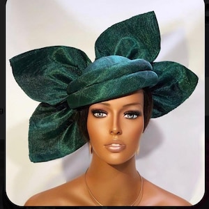 Può includere: Un grande cappello di tessuto verde con un grande fiocco in cima. Il cappello è fatto di un materiale lucido, simile al raso.