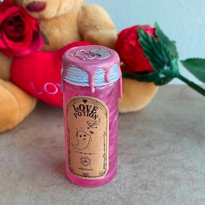 May include: A pink glass bottle with a white and silver wax seal. The bottle is labeled "Love Potion" and features a cartoon ghost with hearts. The bottle is sitting on a gray surface.
