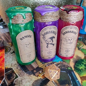 Puede incluir: Tres botellas de poción coloridas etiquetadas como "Ranger", "Sorcerer" y "Barbarian". Cada botella tiene un color, una etiqueta y una cuerda decorativa únicos. Un dado de veinte caras descansa sobre un tablero de juego, sugiriendo un tema de juego de rol.