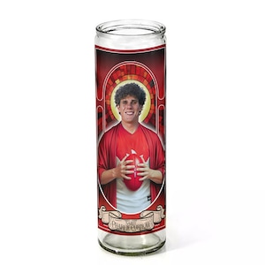 May include: A clear glass candle with a red and white label featuring a portrait of a man holding a football. The label includes the text "Saint Charlie Currow" in a banner design. The man is wearing a red shirt.