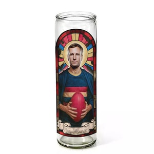 May include: Clear glass prayer candle with a stained-glass style design. It features a portrait of a man holding a red football, wearing a blue and red striped shirt. The text "SAINT JORDAN DAWSON" is printed on a banner.