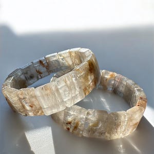 May include: Two clear quartz bracelets with rectangular beads. The bracelets have a mix of clear and light brown tones, with some internal inclusions. The bracelets are displayed on a white surface.
