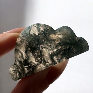 May include: A cloud-shaped, polished stone with a unique pattern of dark green and black inclusions. The stone is held in a hand, showcasing its natural beauty and intricate details. The stone's shape and color make it a unique decorative item.