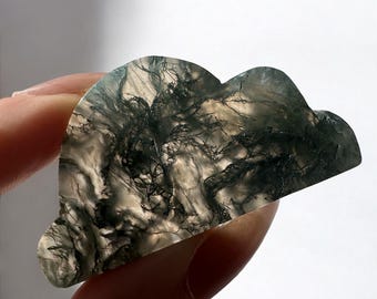 May include: A cloud-shaped, polished stone with a unique pattern of dark green and black inclusions. The stone is held in a hand, showcasing its natural beauty and intricate details. The stone's shape and color make it a unique decorative item.