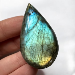 May include: A teardrop-shaped labradorite gemstone with a polished surface. The stone displays a vibrant play of color, with iridescent blues, greens, and yellows. The gemstone is held in a hand against a white background.