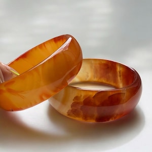 May include: Two translucent orange and amber-colored bangles. The bangles are smooth and glossy, with variations in color and pattern. The light shines through the bangles, highlighting their natural beauty. The bangles are a warm, earthy tone.