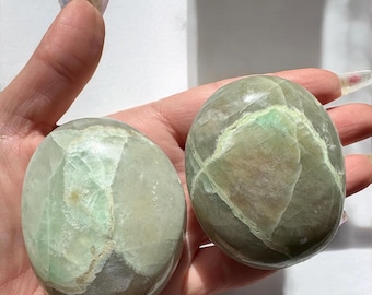 May include: Two green and white polished selenite crystal palm stones. The stones are smooth and have a natural, organic shape.