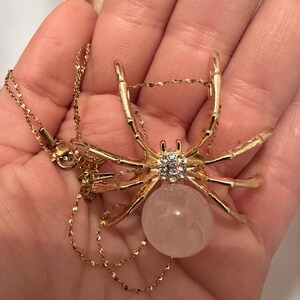 May include: Gold-toned spider pendant necklace with a rose quartz bead body and crystal accents. The spider's legs are outstretched, and the chain is delicate. The necklace is displayed on a hand.