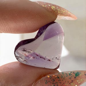 May include: A heart-shaped amethyst gemstone, held between fingers. The gemstone is a translucent purple and white color, with visible inclusions. The edges are smooth and polished. The nails are painted with glitter.