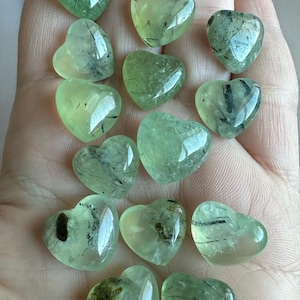 May include: A collection of heart-shaped prehnite stones. The stones are a translucent pale green with dark inclusions, and are polished to a smooth, glossy finish. The hearts vary slightly in size and the inclusions vary in pattern.