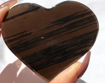 May include: A heart-shaped stone with a polished surface. The stone is a deep brown color with dark, horizontal stripes. The heart is held by a hand with long, polished nails. The background is white.