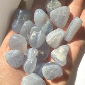 May include: A collection of polished blue lace agate stones in various shapes and sizes. The stones have a pale blue color with white banding and are held in an open hand. The natural gemstones are smooth and appear translucent.