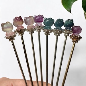 May include: A collection of decorative hair pins. Each pin features a cat-shaped gemstone head in various colors, including pink, purple, and green. The cat heads sit atop silver-colored metal stems with a decorative floral base.