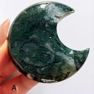 May include: A crescent-shaped, polished stone with a moss agate pattern. The stone features a dark green and white swirling pattern, with a smooth, reflective surface. The crescent is held by a hand, with the letter "A" visible.