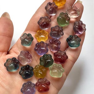 May include: A collection of colorful, paw-shaped, translucent gemstones in various hues, including orange, yellow, green, purple, and red. The gemstones are held in a hand, showcasing their polished surfaces and intricate details. The paw prints are a charming and unique design.