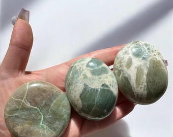 May include: Three green and white patterned polished stones, also known as green aventurine, in various shapes and sizes, held in a hand.