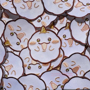 May include: A close-up of a pile of white embroidered patches with brown outlines. Each patch features a cartoon cloud with a smiling face and a pink tongue sticking out. Some of the clouds have a gold horn on top.