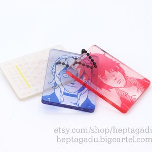 May include: Three acrylic keychains with different images. The keychain on the left is clear with a yellow line and black text. The keychain in the middle is blue with a cartoon image of a person with white hair and blue eyes. The keychain on the right is red with a cartoon image of a person with red hair and brown eyes.