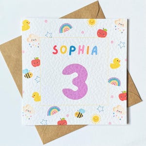 K&ouml;nnte beinhalten: Eine wei&szlig;e Karte mit einer lila Zahl 3 und dem Namen "SOPHIA" in Regenbogenfarben. Die Karte ist mit einem bunten Muster aus Regenb&ouml;gen, Sternen, Wolken, Bienen, &Auml;pfeln und Sonnen verziert.