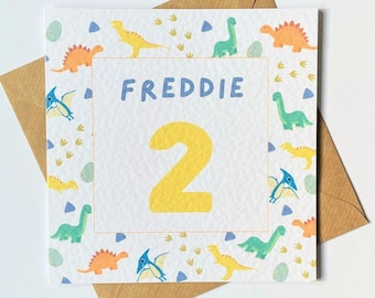 May include: A white card with a dinosaur theme. The card has a yellow number 2 and the name Freddie in blue letters. The card is surrounded by a border of colorful dinosaurs and dinosaur footprints.