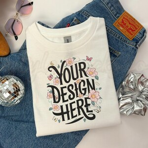 Könnte beinhalten: Weißes T-Shirt mit floralem Design und dem Aufdruck "YOUR DESIGN HERE" auf der Vorderseite. Das T-Shirt ist gefaltet und auf einer blauen Jeans gelegt.