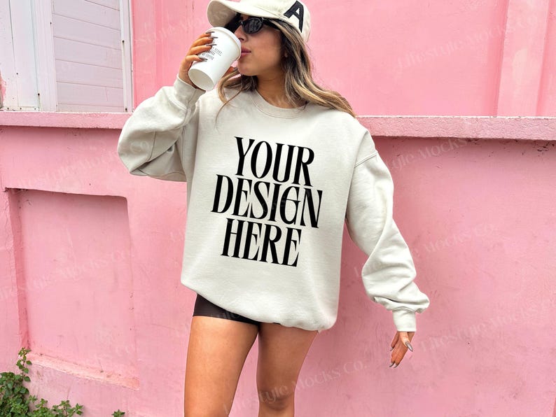 Może przedstawiać: Biała bluza z nadrukiem "YOUR DESIGN HERE" w kolorze czarnym. Modelka nosi bluzę z czarną czapką bejsbolową i okularami przeciwsłonecznymi.