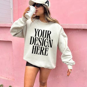 Może przedstawiać: Biała bluza z nadrukiem "YOUR DESIGN HERE" w kolorze czarnym. Modelka nosi bluzę z czarną czapką bejsbolową i okularami przeciwsłonecznymi.