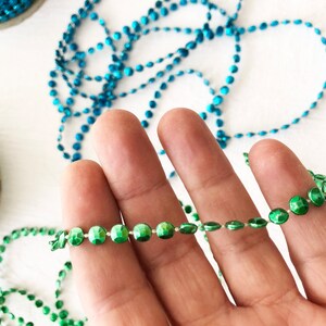 Op de afbeelding: Een streng groene metalen kralen, vastgehouden in een hand, met een spoel groene kralen en een spoel blauwe kralen op de achtergrond.