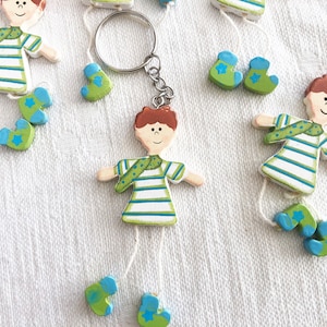 May include: Wooden keychain featuring a cartoon figure with a striped shirt, green scarf, and blue boots. The figure is attached to a silver key ring.