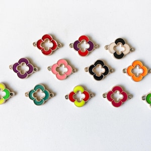 May include: A collection of twelve clover-shaped metal charms featuring colourful enamel fill. Each charm has a central cutout and two small loops on opposite sides. The colours include red, purple, white, black, pink, orange, yellow, green, and magenta.