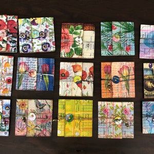 May include: A collection of 12 small, square, handmade journals with various floral and patterned covers. Each journal has a button closure and is tied with twine.