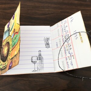 May include: A small, handmade notebook with a colorful cover featuring a jar of olives, a bottle of wine, and a glass of wine. The notebook is open to a page with a handwritten list of numbers and the text "fk 1993".