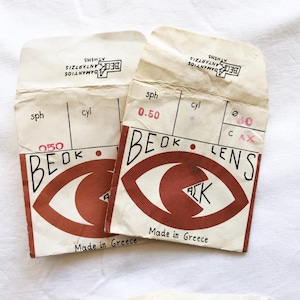 Może przedstawiać: Dwa opakowania soczewek w stylu vintage z brązową grafiką oka i tekstem "BEOK LENS" i "Made in Greece". Opakowania mają odręczne numery i tekst, z marką "BECK ADAMANTIOS ATHENS" na górze.