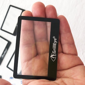 Può includere: Uno schermo di ricambio GrillEye® rettangolare nero tenuto in mano, con diversi altri sullo sfondo. Lo schermo ha un centro trasparente e il logo GrillEye® sul lato.