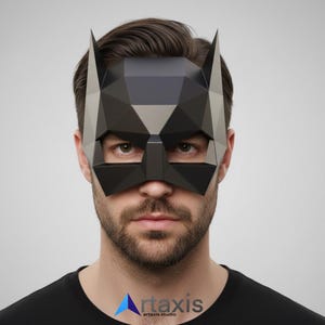 Op de afbeelding: Een zwart geometrisch Batman-masker met puntige oren. Het masker is gemaakt van hoekige, facetvormige vormen en bedekt de ogen en neus. Het masker wordt gedragen door een persoon die een zwart shirt draagt. Het logo van artaxis studio staat linksonder.