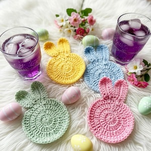 以下が含まれることがあります： 黄色、青、緑、ピンクの、かぎ針編みで作られたウサギ型のコースターのコレクション。紫色の液体と氷が入ったグラス2つ、イースターエッグ、白いふわふわの表面に花が飾られています。