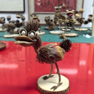 以下が含まれることがあります： 天然素材で作られた、気まぐれな手作りの鳥の彫刻。鳥は、詳細な体、頭、脚を持ち、大きくて丸い目をしています。小さな木の台座に立っており、ユニークで素朴な美学を示しています。他の同様の彫刻が背景にあります。