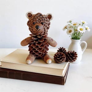 Könnte beinhalten: Eine handgefertigte braune Tannenzapfen-Bärenfigur mit bemalten Details, die auf einem Bücherstapel sitzt. Der Bär hat ein lächelndes Gesicht und ist von Tannenzapfen und einer kleinen weißen Vase mit Gänseblümchen umgeben.