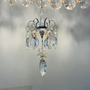 May include: A small, ornate chandelier with multiple clear crystal pendants and accents. The chandelier has a silver frame and is suspended from a chain. The top of the chandelier has a gold-colored frame with large, clear crystal accents.