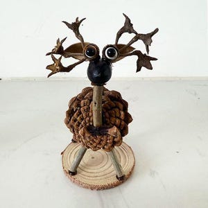 Puede incluir: Una figura de ciervo hecha a mano y caprichosa, elaborada con materiales naturales. El ciervo presenta un cuerpo de piña, patas de ramitas y una cabeza hecha de una semilla negra y otros elementos naturales. Los ojos son blancos con pupilas negras. Se apoya sobre una pequeña base de madera.