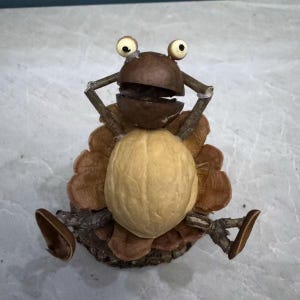Puede incluir: Una escultura caprichosa de rana hecha con materiales naturales. La rana presenta un cuerpo de nuez marrón claro, una cabeza de cáscara de nuez marrón oscuro y brazos de ramitas. Los ojos son pequeños, redondos y blancos con pupilas negras. La rana está sentada sobre un lecho de escamas de piña marrones.