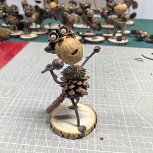 May include: A handcrafted sculpture of a creature made from natural materials. The head is a walnut with googly eyes, the body is a pinecone, and the arms are twigs. It stands on a wooden disc base. Other similar sculptures are in the background.