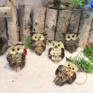 Puede incluir: Cinco figuras decorativas de búhos. Cada búho está hecho de materiales naturales, incluyendo piñas, ramitas y paja. Los búhos tienen ojos grandes y redondos y están colocados sobre una superficie blanca, con un fondo de troncos de abedul y vegetación.