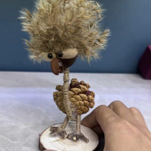 Peut inclure: Une figurine d'oiseau fantaisiste faite à la main à partir de matériaux naturels. L'oiseau présente une tête duveteuse et beige, un corps en forme de graine et des pattes en brindilles, le tout monté sur une petite tranche de bois. Le bec et les yeux sont faits de petites graines brun foncé.