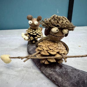 Puede incluir: Dos figuras caprichosas hechas de piñas, nueces y ramitas. Una figura sostiene una ramita con una nuez, mientras que la otra tiene una apariencia de oso. Las figuras están sobre una superficie gris texturizada, creando una estética lúdica y natural.